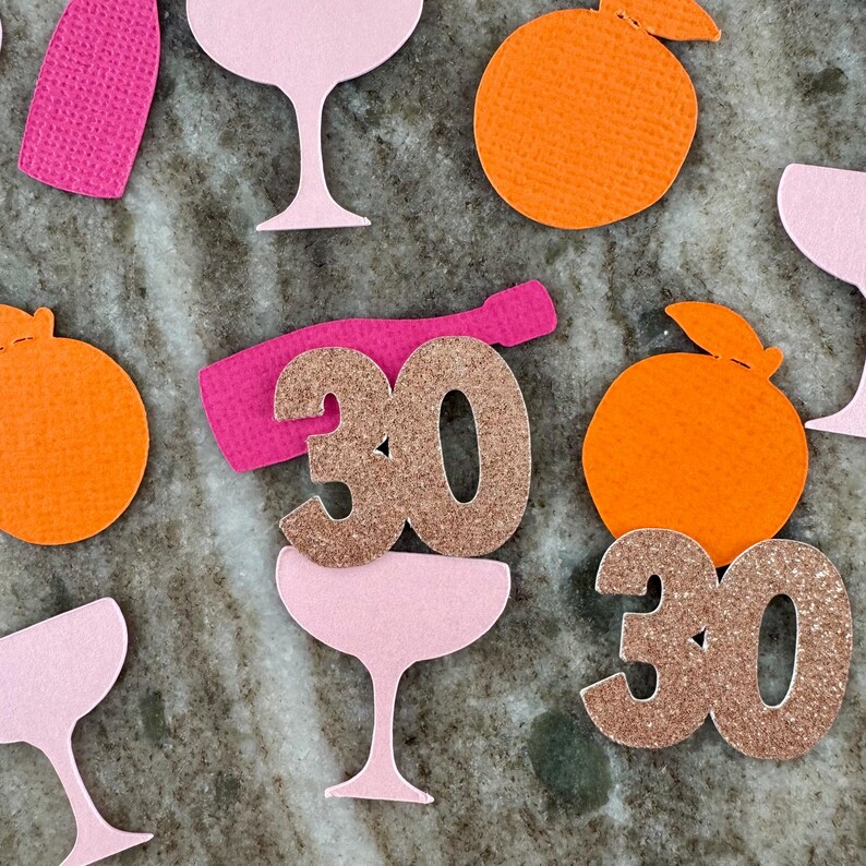 May include: A collection of party-themed paper cutouts, including champagne glasses, champagne bottles, and orange slices. Two glittery gold "30" cutouts are featured, suggesting a milestone birthday celebration. The color palette includes pink, orange, and gold.
