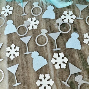 May include: Assortment of light blue and white paper confetti featuring snowflakes, martini glasses, rings, and winter hats.