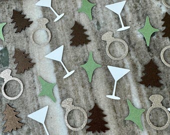 May include: Assorted confetti pieces scattered on a surface. The confetti includes white martini glasses, green star shapes, brown Christmas trees, and glittery engagement rings. The items are arranged randomly, creating a festive and celebratory feel.