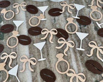 May include: Assorted confetti pieces scattered on a surface. The confetti includes engagement rings, martini glasses, coffee beans, and bows in shades of gold, white, and brown. The background features a sheer, white fabric.