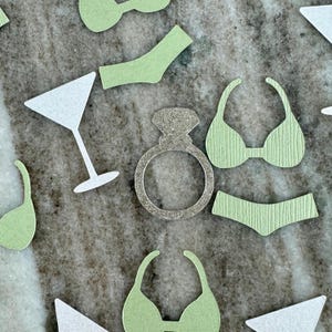 May include: A set of green and white paper cutouts for a bachelorette party. The cutouts include a ring, a martini glass, and a bikini.