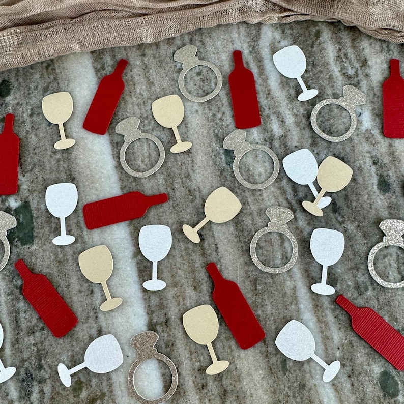May include: Assortment of confetti featuring wine glasses, wine bottles, and engagement rings in red, white, and gold glitter.