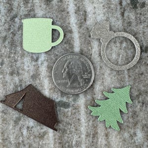 May include: A set of five paper cutouts: a green camping mug, a silver ring, a brown tent, a green pine tree, and a silver quarter with the words "United States of America" and "Liberty" printed on it.