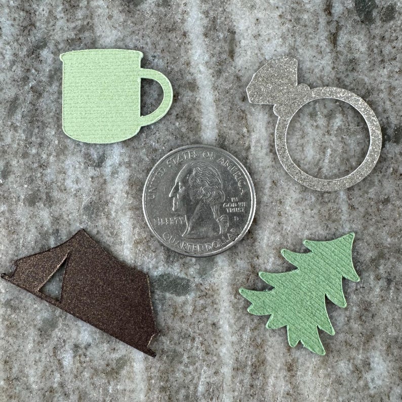May include: A set of five paper cutouts: a green camping mug, a silver ring, a brown tent, a green pine tree, and a silver quarter with the words "United States of America" and "Liberty" printed on it.
