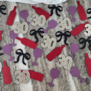 May include: A collection of party decorations featuring red wine bottles, purple wine glasses, black bows, and glittery silver number 30 cutouts. The items are scattered on a textured surface, suitable for a 30th birthday celebration.