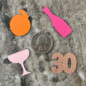 May include: An assortment of party-themed paper cutouts, including an orange, a pink champagne bottle, a pink cocktail glass, and the number 30 in glittery copper. A U.S. quarter is included for size reference.