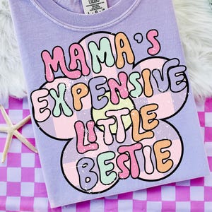 May include: A light purple t-shirt with the text "MAMA'S EXPENSIVE LITTLE BESTIE" in a colorful, retro font. The text is set against a flower-shaped design with a checkered pattern. The shirt is on a pink and purple checkered surface.