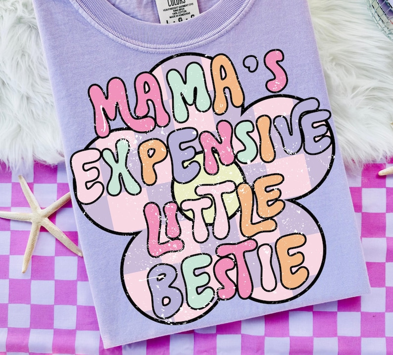 May include: A light purple t-shirt with the text "MAMA'S EXPENSIVE LITTLE BESTIE" in a colorful, retro font. The text is set against a flower-shaped design with a checkered pattern. The shirt is on a pink and purple checkered surface.