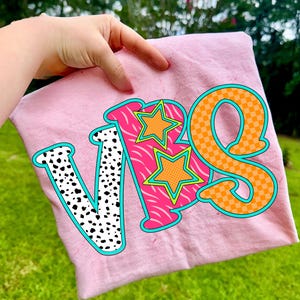 May include: A light pink t-shirt with the letters "VBS" in a colorful design. The letter "V" is white with black spots, the "B" is pink with a zebra pattern, and the "S" is orange with a checkered pattern. Two orange stars are on the "B".
