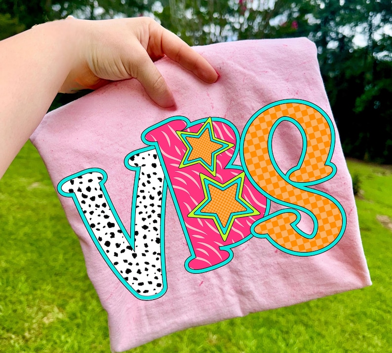 May include: A light pink t-shirt with the letters "VBS" in a colorful design. The letter "V" is white with black spots, the "B" is pink with a zebra pattern, and the "S" is orange with a checkered pattern. Two orange stars are on the "B".