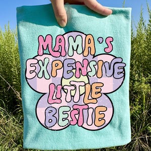 May include: A light blue t-shirt with the text "MAMA'S EXPENSIVE LITTLE BESTIE" in a colorful, retro font. The text is set against a flower-shaped design with a checkered pattern.