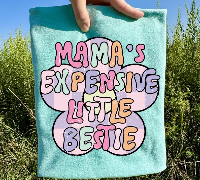 May include: A light blue t-shirt with the text "MAMA'S EXPENSIVE LITTLE BESTIE" in a colorful, retro font. The text is set against a flower-shaped design with a checkered pattern.