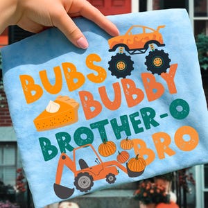 May include: Light blue t-shirt with the words "Bubs, Bubby, Brother-O, Bro" in orange and green. The shirt features illustrations of a monster truck, a slice of pumpkin pie, pumpkins, and a construction vehicle.