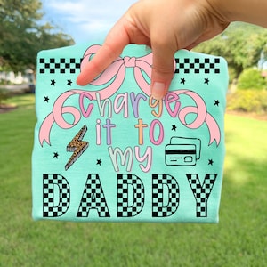 Puede incluir: Camiseta de color verde azulado claro con la frase "Charge it to my Daddy" en letras coloridas. El diseño incluye un lazo rosa, un rayo con estampado de leopardo e ilustraciones de tarjetas de crédito.