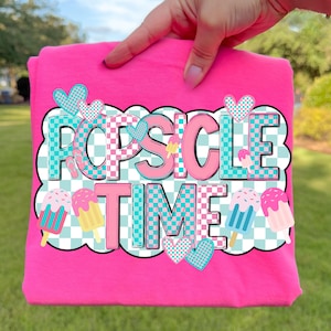 May include: A bright pink t-shirt with the words "POPSICLE TIME" in a colorful, playful font. The design includes popsicle illustrations and heart accents, set against a checkered background. The shirt is held up by a hand.