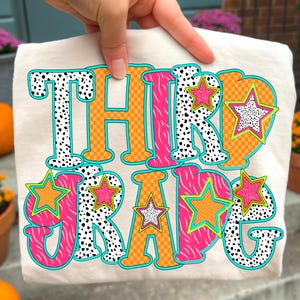May include: Cream-colored t-shirt with the words "THIRD GRADE" in a colorful, playful design. The text features orange, pink, and black and white patterns, with star accents. The shirt is held up by a hand.