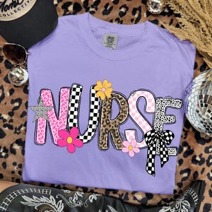 May include: A lilac-colored t-shirt with the word "NURSE" in a playful, patterned design. The letters feature pink leopard print, checkered, and floral accents. A black hat and sunglasses are visible in the background.