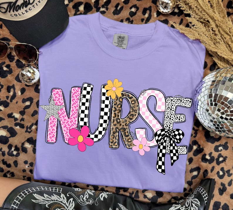 May include: A lilac-colored t-shirt with the word "NURSE" in a playful, patterned design. The letters feature pink leopard print, checkered, and floral accents. A black hat and sunglasses are visible in the background.