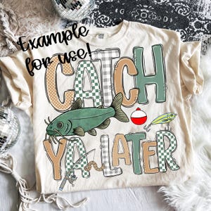May include: Cream-colored t-shirt with the phrase "CATCH YA LATER" in a mix of fonts and patterns. The design includes a green catfish, fishing lures, and a bobber. The shirt is a Comfort Colors brand.