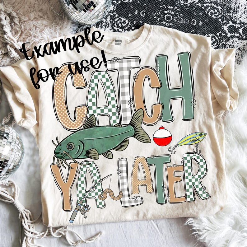 May include: Cream-colored t-shirt with the phrase "CATCH YA LATER" in a mix of fonts and patterns. The design includes a green catfish, fishing lures, and a bobber. The shirt is a Comfort Colors brand.
