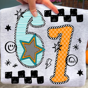 May include: A light grey sweatshirt displaying the numbers '67' in a vibrant, playful style. The number '6' is turquoise with a star, and the number '7' is orange. Black and white checkered patterns and doodle-style stars and smiley faces complete the design.