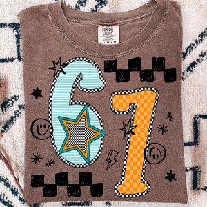 May include: Brown Comfort Colors t-shirt featuring the number "67" in a graphic design. The "6" is light blue with white stripes and a star. The "7" is orange with a checkered pattern. Black stars, smiley faces, and lightning bolts surround the numbers.