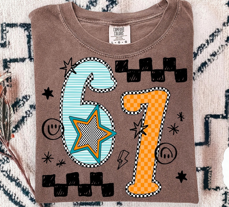 May include: Brown Comfort Colors t-shirt featuring the number "67" in a graphic design. The "6" is light blue with white stripes and a star. The "7" is orange with a checkered pattern. Black stars, smiley faces, and lightning bolts surround the numbers.