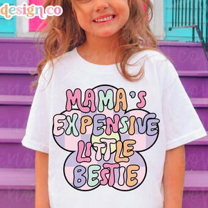 May include: White t-shirt with the text "MAMA'S EXPENSIVE LITTLE BESTIE" in a colorful, retro flower design. The shirt is worn by a young girl. The brand name "ZTL design co" is in the top left corner.