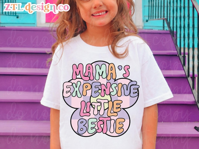 May include: White t-shirt with the text "MAMA'S EXPENSIVE LITTLE BESTIE" in a colorful, retro flower design. The shirt is worn by a young girl. The brand name "ZTL design co" is in the top left corner.