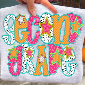 May include: A gray sweatshirt with the words "SECOND GRADE" in a colorful, playful design. The text features orange, pink, and black patterns, with star accents. The design is outlined in teal.