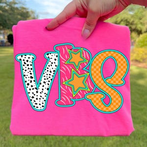 May include: A bright pink t-shirt with the letters "VBS" in a colorful design. The "V" is white with black spots, the "B" has a zebra print, and the "S" is orange with a checkered pattern. Two orange stars with green outlines are also featured.