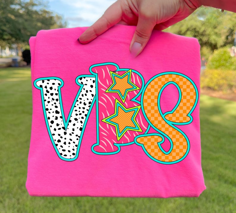 May include: A bright pink t-shirt with the letters "VBS" in a colorful design. The "V" is white with black spots, the "B" has a zebra print, and the "S" is orange with a checkered pattern. Two orange stars with green outlines are also featured.