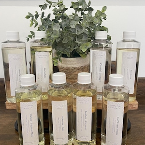 May include: Ten clear glass bottles with white lids and labels. The bottles contain reed diffusers with various scents, including Seaside Linen, Waterfall, Coastal Cottage, Sunshine, Mango + Coconut Milk, Oakmoss + Sage, Bamboo + Coconut, and Gulf Coast Breeze. The bottles are arranged in two rows on a wooden shelf.