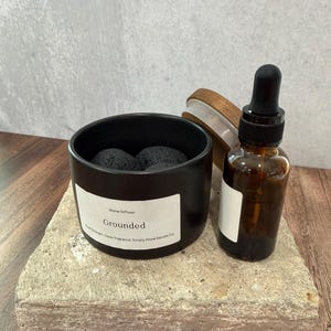 May include: A black stone diffuser with a white label reading Stone Diffuser Grounded, containing dark porous stones. Beside it sits an amber glass dropper bottle with a black cap and a wooden lid with a white silicone seal.