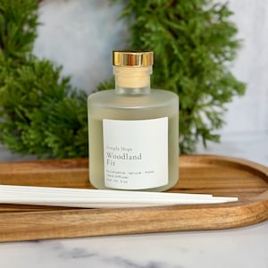 May include: A frosted glass reed diffuser bottle with a gold cap and a white label reading Simply Hope Woodland Fir eucalyptus spruce moss reed diffuser Net Wt. 5 oz, accompanied by white reed sticks on a wooden tray.