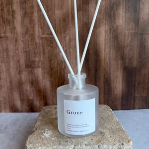 May include: A frosted glass reed diffuser bottle with three white reeds. The white label reads Grove, cinnamon/berry/pecan, hand poured reed diffuser, 5 oz., Simply Hope Candle Co. The bottle has a cylindrical shape and a clear glass neck.