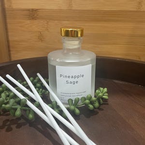 May include: A frosted glass reed diffuser with a gold cap, labeled "Pineapple Sage." White reeds and green leaves are arranged around the bottle. The diffuser is on a wooden tray, suggesting a home fragrance product.