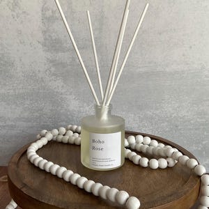 May include: A frosted glass reed diffuser with white reeds and a label that reads "Boho Rose." The diffuser sits on a wooden tray, surrounded by a white beaded garland. The background is a textured gray wall.