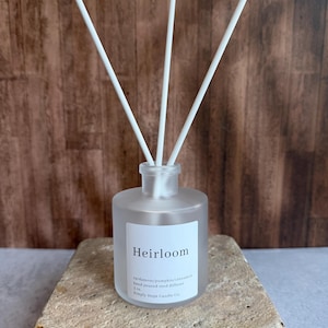 May include: A frosted glass reed diffuser bottle with three white reeds. The white label reads Heirloom, cardamom/pumpkin/cinnamon, hand poured reed diffuser, 2 oz., Simply Hope Candle Co. The bottle has a cylindrical shape and a clear glass neck.