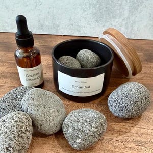May include: Aromatherapy set featuring a black stone diffuser with two gray stones inside, a bottle of Grounded Diffuser Oil, and several gray stones. The diffuser has a wooden lid. The label on the diffuser reads "Grounded".