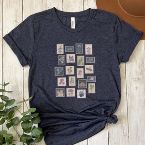 May include: A dark blue t-shirt with a graphic print of a collection of vintage postage stamps featuring flowers, insects, and mushrooms.