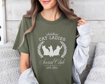 May include: A green t-shirt with the text "childless CAT LADIES Social Club EST. 2021" and a graphic of three cats in a wreath.