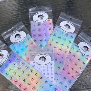 May include: Clear plastic bags containing iridescent fishing line weight labels. Each label displays a number indicating the line weight, ranging from 2 to 80, in pounds. The labels also feature a logo with a fish.