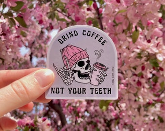 Op de afbeelding: Een witte sticker met een roze schedel die een roze muts draagt en een koffiebeker vasthoudt. De sticker zegt "Grind Coffee Not Your Teeth" met de tekst "Perfectly White Prints" in kleiner lettertype.