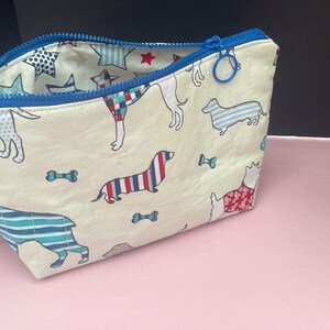 May include: A cream-colored zippered pouch with a blue zipper and a pattern of colorful dogs on a white background. The pouch is open and the inside lining is blue with white stars.