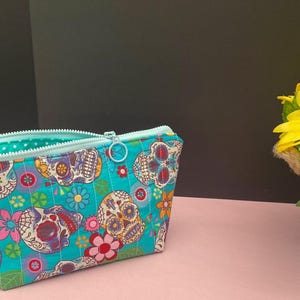 May include: A turquoise zippered pouch with a colorful sugar skull pattern. The pouch features a light blue zipper and a matching interior. A small tag is attached to the side. A pot of sunflowers is to the right.