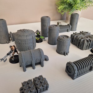 May include: A collection of gray, 3D printed, sci-fi themed miniature buildings. The buildings are various shapes and sizes, including cylinders, domes, and rectangular structures. Some of the buildings have pipes and other details.