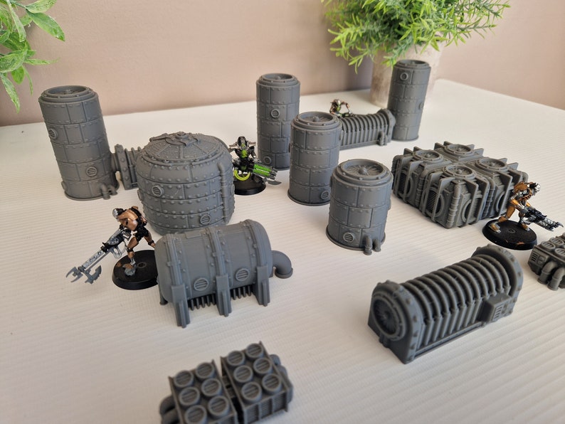 May include: A collection of gray, 3D printed, sci-fi themed miniature buildings. The buildings are various shapes and sizes, including cylinders, domes, and rectangular structures. Some of the buildings have pipes and other details.