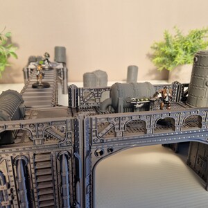 May include: A gray, metal, modular, sci-fi themed terrain set for tabletop gaming. The set features a bridge, a walkway, and a variety of pipes and other industrial details. The set is painted in a dark gray color and has a weathered look.