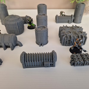 May include: A collection of gray 3D printed miniature buildings and figures. The buildings are various shapes and sizes, including cylinders, rectangular boxes, and a square building with four round vents. There are also two miniature figures, one green and one gold.